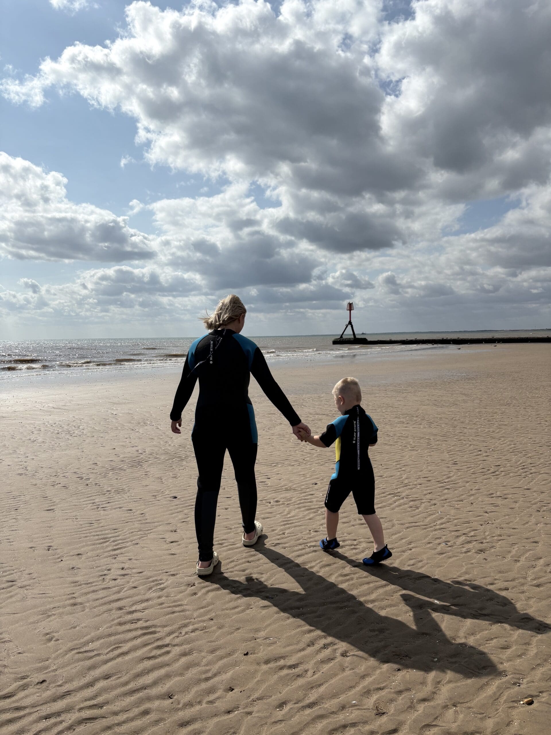A woman and child in wetsuits walk hand in hand on a sandy beach under a cloudy sky. The ocean waves are gentle, creating a serene, bonding moment.